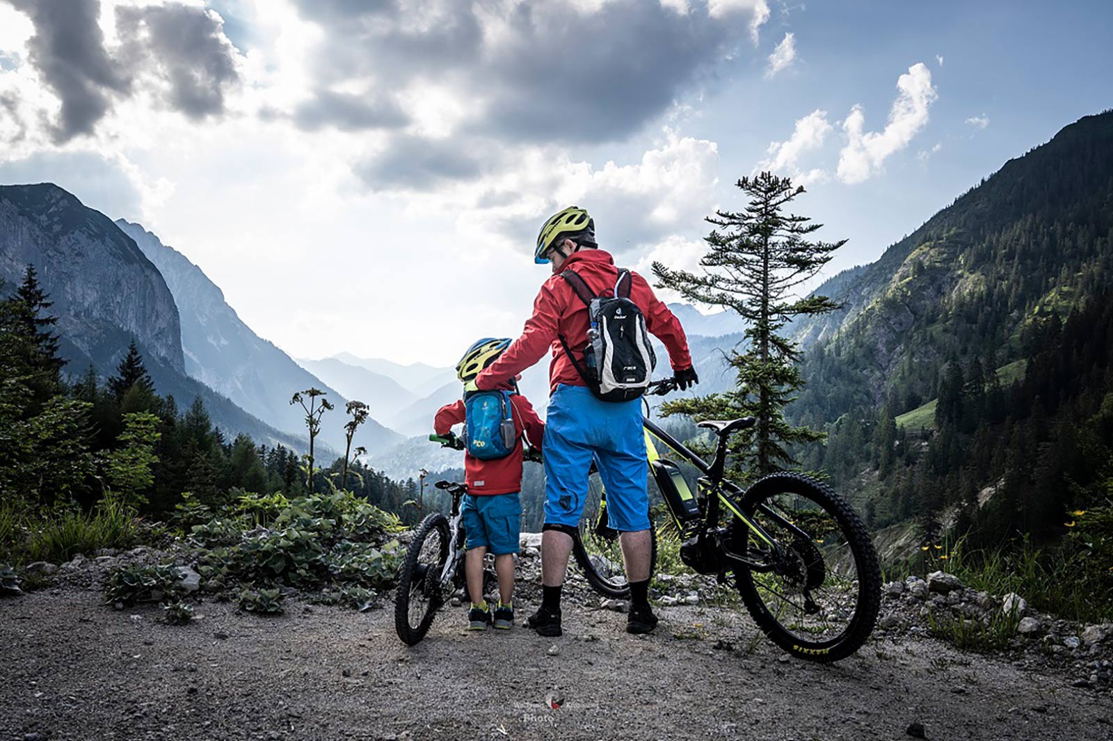 Ein Kind und ein Erwachsener mit Mountainbikes jeweils mit blauen Hosen und roten Jacken vor einer Bergkulisse.